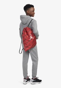 Crvena torba s rastezom i crnim natpisom "JORDAN" te bijelim logotipom; nosi je dijete u sivoj dukserici i trenirkama, uparena s crnim tenisicama.