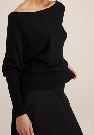 Kvinde iført en løs sort off-shoulder sweater og matchende sort nederdel mod en beige baggrund.