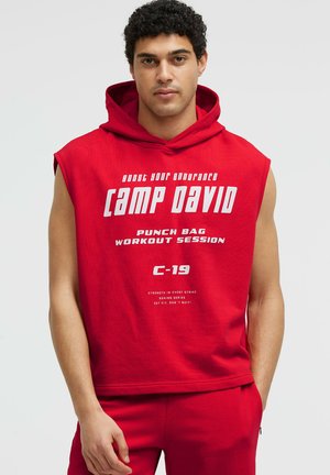 Young man wearing a red sleeveless hoodie with white text about boxing and endurance, standing against a plain light background.