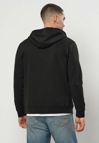 Melna džemperis ar plašu kapuci, izgatavots no mīksta auduma. Aprīkots ar ribotām piedurknēm un apakšmalu, valkāts virs baltas krekla, saderināts ar gaiši zilām džinsām.