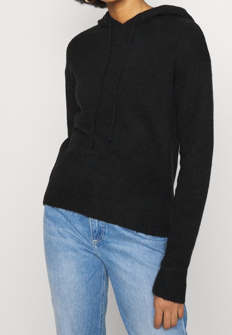 Femme portant un sweat à capuche noir avec cordons et un jean bleu clair, debout devant un fond clair uni.