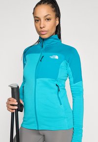 Ελαφρύ, πετρόλ μπουφάν με φερμουάρ, υφασμένο με υφή, αντίθετα πάνελ, δύο τσέπες με φερμουάρ και ψηλό γιακά. Συνοδεύεται από μπαστούνια πεζοπορίας.