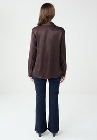 Ciemnobrązowa jedwabna koszula z długim rękawem, z wydłużonym kołnierzykiem i mankietami na guziki, w połączeniu z ciemnymi dzwonami jeansami. Gładka tekstura i dopasowany krój.