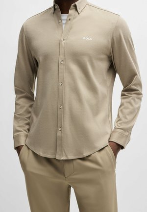 Homme portant une chemise beige à manches longues boutonnée avec le logo "BOSS" sur la poitrine et un pantalon beige assorti, mains dans les poches.