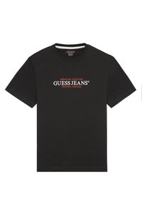 Musta puuvillane T-särk, millel on ümar kaelus, lühikesed varrukad ja ees valge ja punase graafiline tekst: "GUESS JEANS AMERICAN TRADITION."