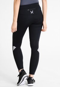 Černé sportovní legíny s hladkým povrchem, které mají reflexní stříbrné detaily na lýtkách a malou zadní kapsu se zipem.