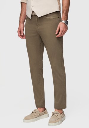 Mand iført olivengrønne slimfit buksere, beige afslappede snørebåndssko og en beige skjorte med et sølv armbåndsur.