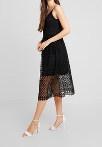 Svart spetsklänning med figursydd överdel och lager-på-lager kjol. Den genomskinliga underdelen avslöjar huden. Bärs med vita klacksandaler med ankelremmar.