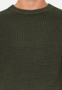Maglione lavorato a maglia di colore verde scuro, con un motivo testurizzato, scollo a girocollo e bordi a costine. Il tessuto appare morbido e spesso.