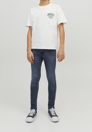 Niño con camiseta blanca de manga corta con el logotipo "Originals SLC", jeans ajustados azul oscuro y zapatillas negras altas con cordones blancos.