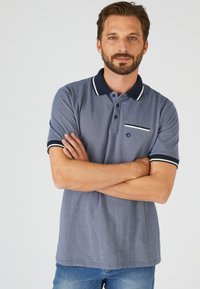 Polo di colore blu navy con finitura testurizzata, bordi bianchi su colletto e maniche, e una tasca sul petto con una striscia orizzontale.