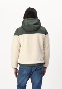 Giacca in pile con cappuccio isolato verde e corpo crema. Presenta un orlo regolabile e polsini elasticizzati. La vista posteriore mostra la texture morbida e il contrasto di colore.