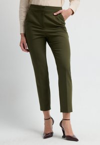 Olijfgroene op maat gemaakte broek met een hoge taille, taps toelopende pijpen en voornaad. Gecombineerd met bordeauxrode puntige hakken en een lichte blouse.