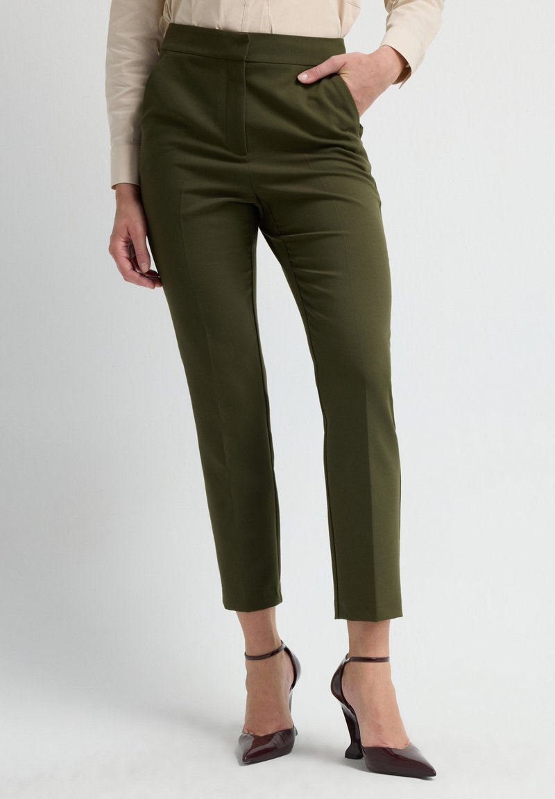 Olijfgroene op maat gemaakte broek met een hoge taille, taps toelopende pijpen en voornaad. Gecombineerd met bordeauxrode puntige hakken en een lichte blouse.