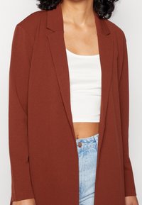 Blazer v rezavé barvě s uvolněným střihem, dlouhými rukávy a otevřeným předním designem. V páru s bílým crop topem a světle modrými džíny s vysokým pasem.
