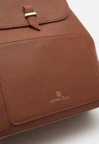 Hnědá texturovaná veganská kožená taška na rameno s uzávěrem na klopu, zlatými akcenty a přední kapsou. Logo značky je vyraženo na dolní části přední strany.