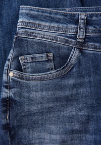 Jean en denim bleu foncé avec une finition texturée, présentant une poche avant et des coutures visibles le long des coutures. Design classique à cinq poches.