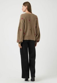 Maglione marrone a maglia con maniche oversize, decorato con ricami floreali rosa. Indossato con pantaloni neri a gamba larga e stivali con tacco.