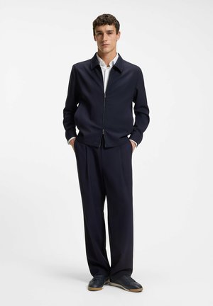 Jeune homme portant une chemise blanche, une veste zippée bleu marine, un pantalon large bleu marine et des baskets foncées, debout les mains dans les poches sur fond blanc.