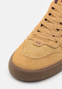 Tan mockasiner i mocka med en kordflorstoffbox, texturerad brun gummisula och matchande snören. Har en liten lila logotyp på plösen.