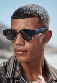 Ochelari de soare negri cu un design unghiular unic, accente metalice și lentile întunecate, purtați de o persoană cu păr scurt și o cămașă cu model.
