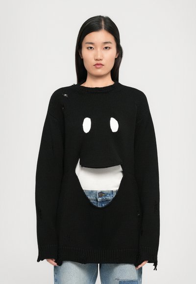 Ung kvinde med langt mørkt hår iført en oversized sort strikket sweater med slidte huller over en hvid top og lyseblå jeans.