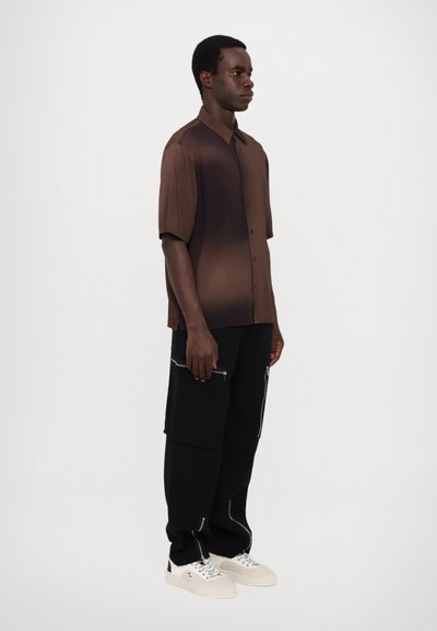 Rövid ujjú, gombos ing átmeneti barna árnyalatban, fekete nadrággal, cipzárokkal, fehér sportcipővel párosítva, egyszerű háttér előtt.