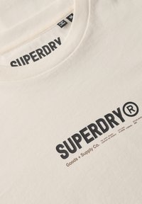 Camiseta de manga corta color crema con el texto negro "SUPERDRY", cuello redondo acanalado y detalles de marca impresos en la tela.
