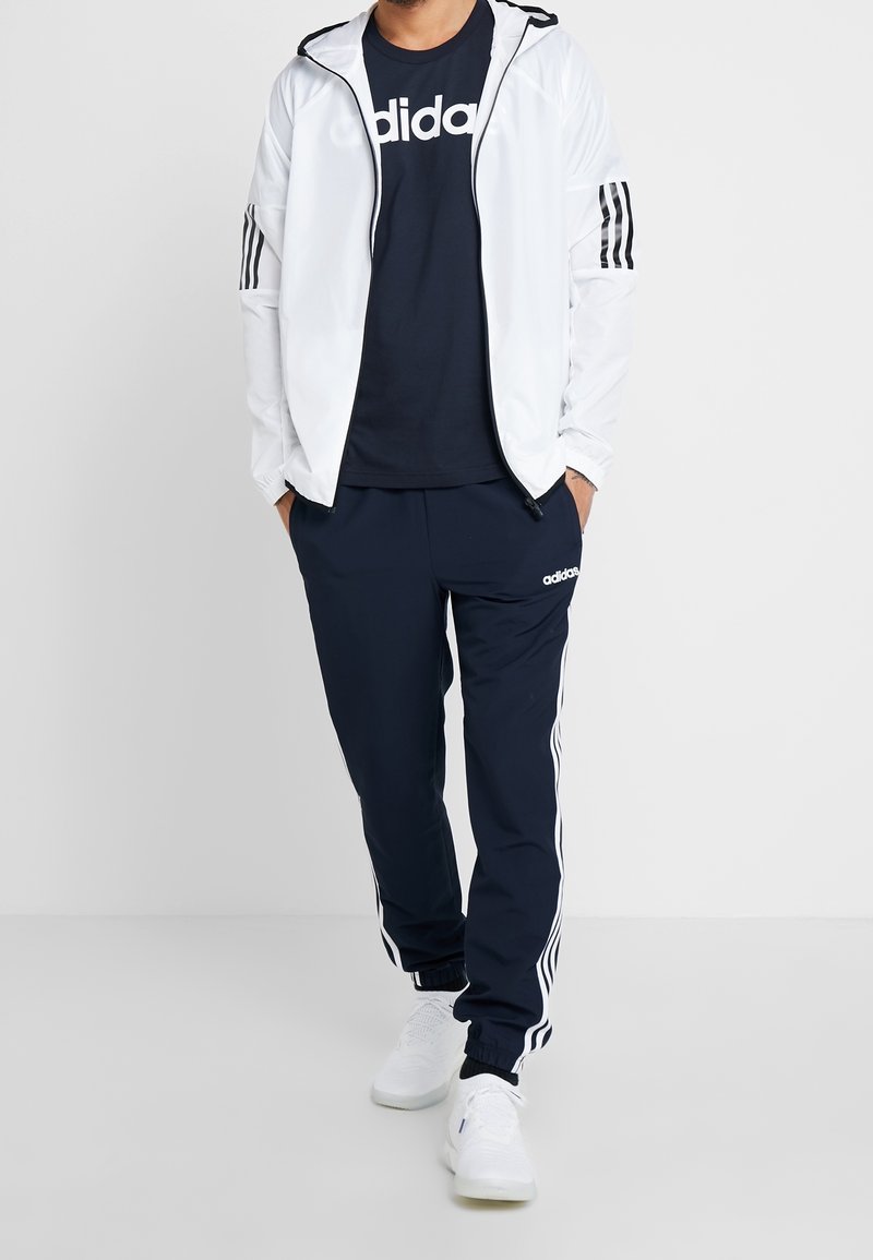 Chaqueta ligera blanca, camiseta de rayas marinas con logo blanco, pantalones de chándal marinos con rayas blancas y zapatillas deportivas blancas. Tejido suave, ajuste casual.