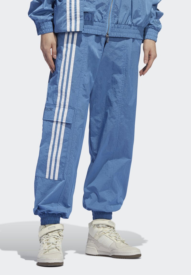 Blauwe trainingsbroek met witte drievoudige strepen en cargozak, gedragen met een bijpassende jas en off-white hoge sneakers op een witte achtergrond.