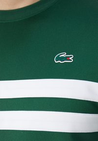 Grön t-shirt med korta ärmar som har två horisontella vita ränder och en liten krokodillogo i grönt och rött på bröstet.