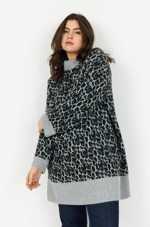 Femme portant un pull léopard gris et noir avec des poignets et un ourlet côtelés, posant devant un mur blanc.