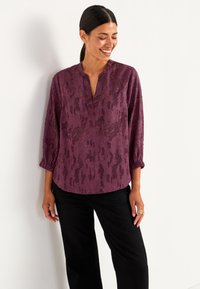 Doorschijnende paarse blouse met een textuurrijk bloemenpatroon, V-hals en driekwart mouwen, gecombineerd met zwarte broek.