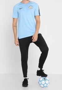 Helles blaues Nike-Trikot mit kurzen Ärmeln und dem Wappen von Manchester City, kombiniert mit schwarzen Sporthosen und schwarzen Fußballschuhen, stehend auf einem Fußball.