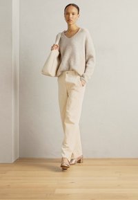 Beige neulepaita V-pääntiellä, kerma-colored housut ja metalliset korkokenkä sandaalit. Pitelee yhdistelevää käsilaukkua. Puulattia, neutraali tausta.