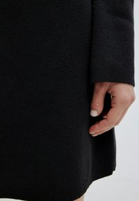 Zwarte gebreide jurk met een gestructureerd oppervlak, lange mouwen en een losse pasvorm, met een hand die nabij de zoom rust.