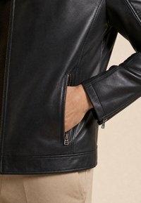 Nahaufnahme einer Person, die eine schwarze Lederjacke trägt, mit der Hand in der seitlich mit Reißverschluss verschlossenen Tasche, unten beige Hose sichtbar.