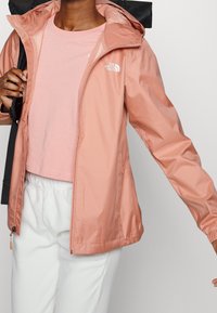 Chaqueta impermeable ligera de color durazno con cierre frontal de cremallera, capucha ajustable y puños elásticos. Se lleva sobre un top rosa y pantalones blancos.