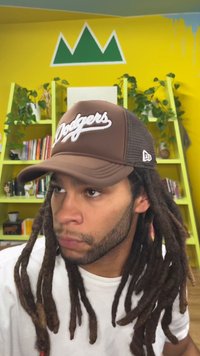 Hnědá baseballová čepice s logem „Dodgers“, síťovanými boky a zakřiveným kšiltem. Osoba s dlouhými tmavými dredy, oblečená v bílém tričku, v barevné místnosti.