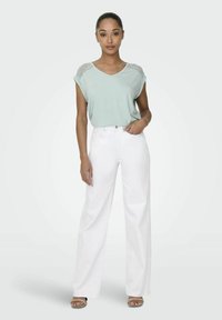 Jasnozielona bluzka z krótkim rękawem, ozdobiona koronką na ramionach, zestawiona z białymi spodniami o wysokim stanie i szerokimi nogawkami oraz beżowymi sandałami na obcasie.