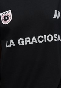 Μαύρο πουκάμισο με λευκό κείμενο "LA GRACIOSA", ροζ και λευκή ασπίδα με μαύρο σχέδιο ήλιου και δύο λευκές κάθετες ρίγες.
