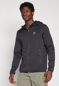 Felpa nera con zip realizzata in tessuto liscio. Presenta un cappuccio, tasche laterali e un piccolo patch con logo sul petto. Cuciture decorative visibili.