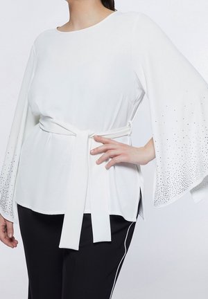 Femme portant un haut blanc ceinturé aux manches larges ornées de petits strass et un pantalon noir avec des bandes blanches latérales.