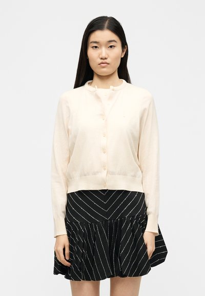 Νεαρή γυναίκα με μακριά σκούρα μαλλιά φοράει ένα κρεμ ζακέτα με κουμπιά και μια μαύρη φούστα με λευκές διαγώνιες ρίγες, στέκεται μπροστά σε ένα μονόχρωμο φόντο.