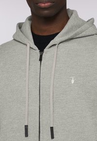 Grå zip-up hoodie i mjukt tyg med svart dragkedja och kontrasterande snören. Har en liten broderad logotyp på bröstet.