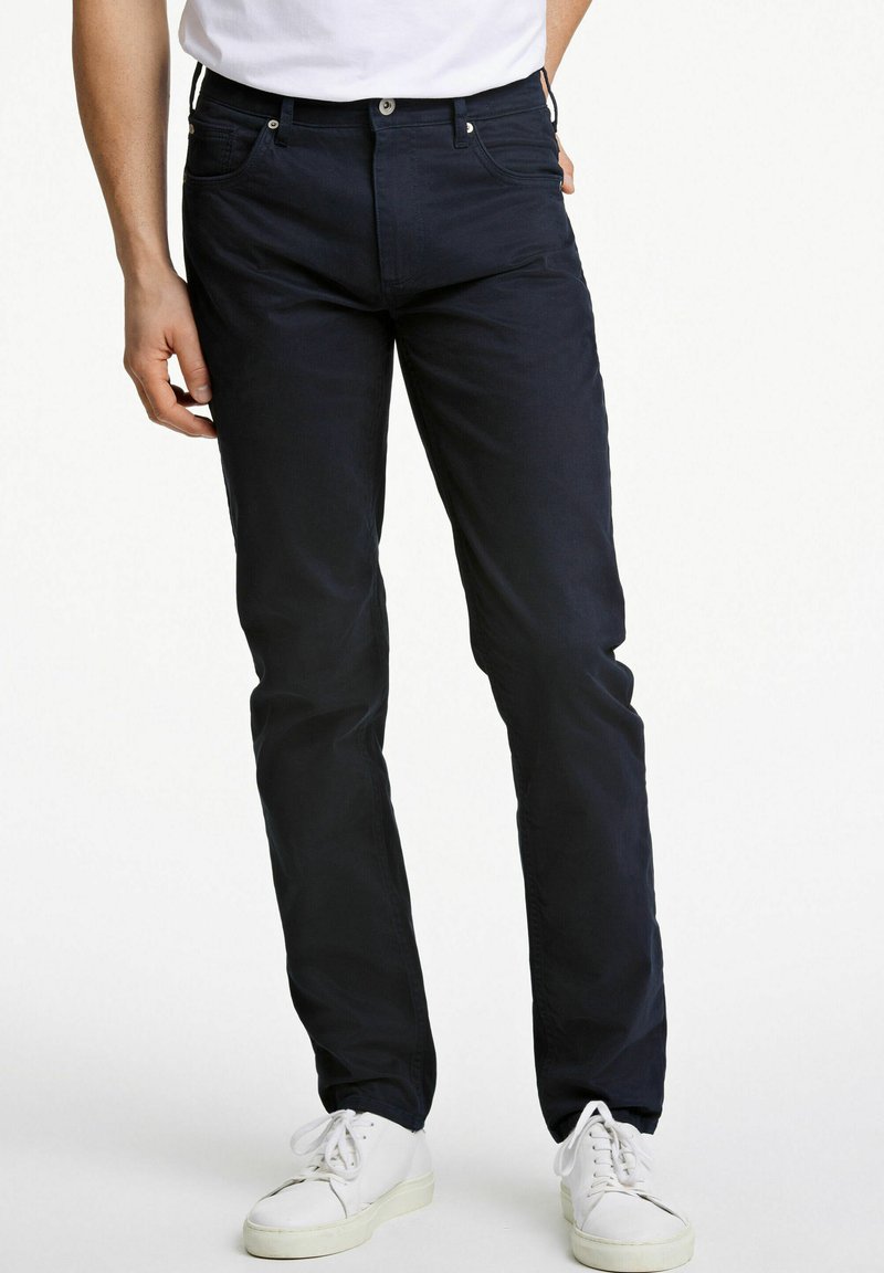Pantaloni in cotone blu navy con vestibilità dritta, dotati di tasche frontali, chiusura con zip e bottone. Indossati con sneakers bianche.