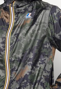 Veste à motif camouflage avec une fermeture éclair rayée verticale et un logo K-Way bleu, rouge et blanc sur la poitrine.