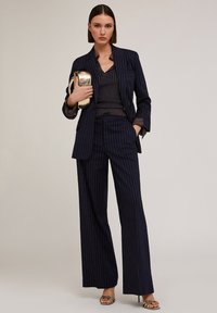 Marineblauw gestreept pak met wijde broekspijpen en een op maat gemaakte blazer. Daaronder een donker patroon overhemd. Een gouden clutch en hakken maken de look compleet.