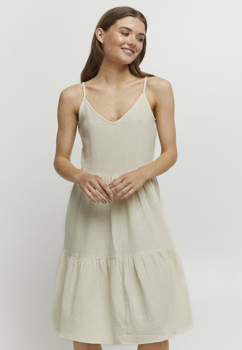 Helles beiges Kleid mit V-Ausschnitt, dünnen Trägern und einem gestuften Rock. Aus strukturiertem Stoff gefertigt, mit dezenten horizontalen Streifen.