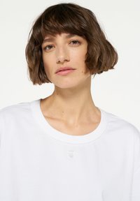 T-shirt bianca a maniche corte in cotone, con scollatura rotonda e un piccolo dettaglio con logo vicino al colletto. Tessuto liscio, vestibilità casual.